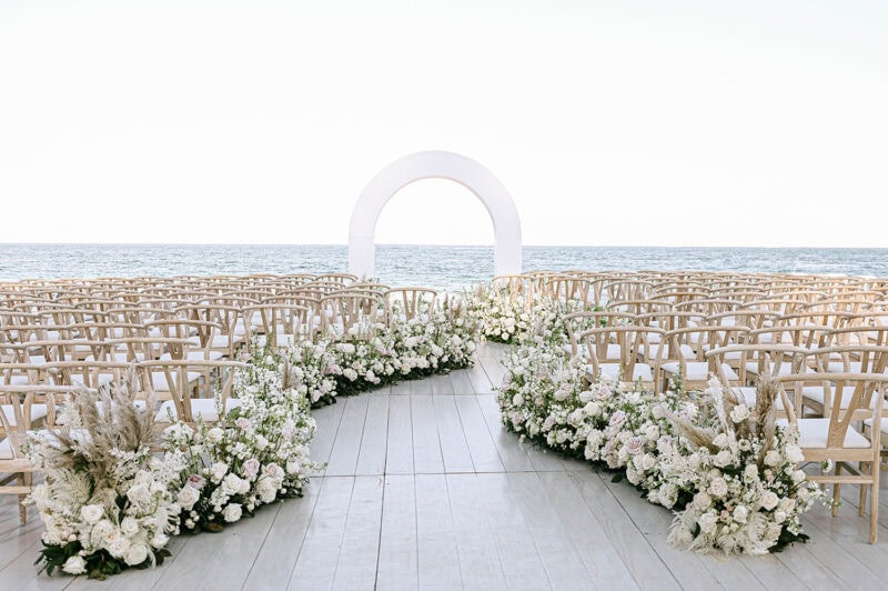 Beach wedding aisle with white flowers and wooden chairs facing the ocean.
