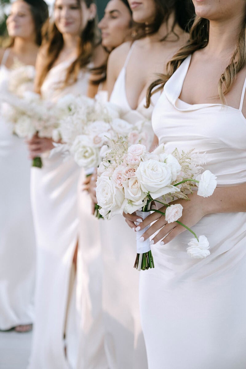 Elegant bridal party with white dresses and floral bouquets at outdoor wedding.
