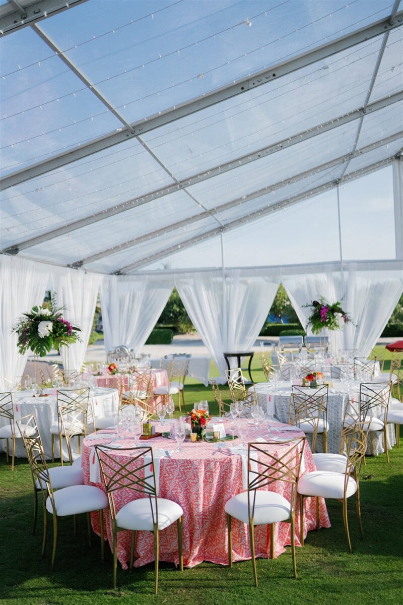 Elegant wedding reception setup inside a clear tent with draped white curtains and floral centerpiec.