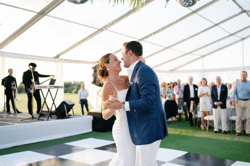 Elegant couple dancing at outdoor wedding reception under a glass tent.