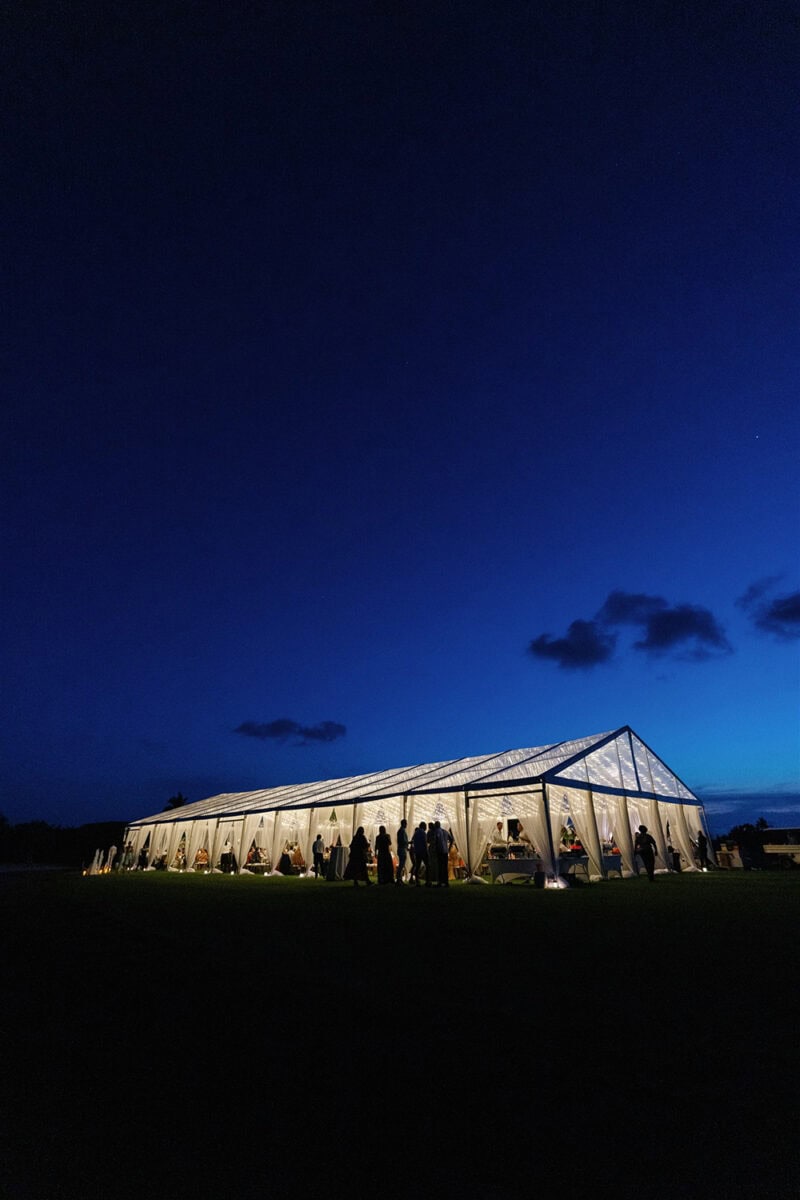 Outdoor wedding reception tent at dusk with guests and string lights.