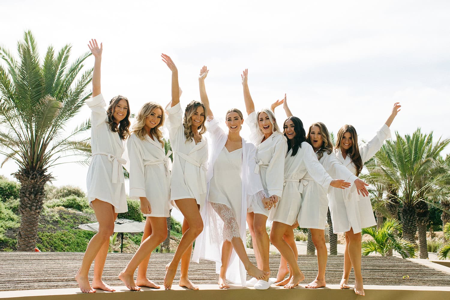 Bridal party in white robes enjoying a joyful moment outdoors with palm trees in the background.