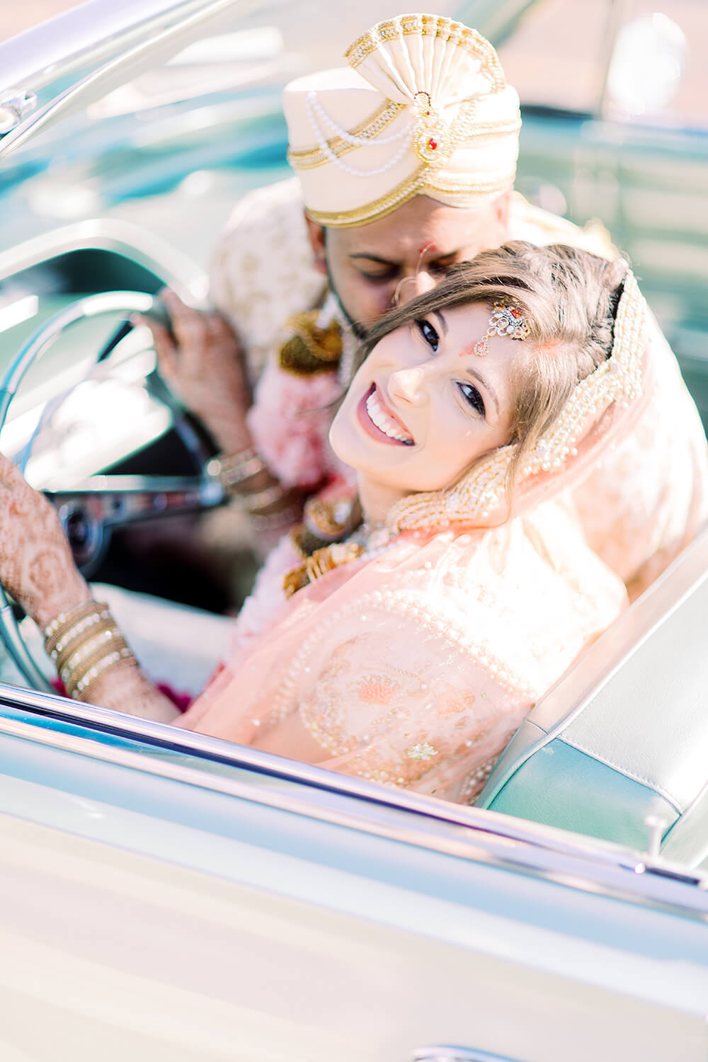 Bride and groom in traditional Indian wedding attire smiling in a classic car.