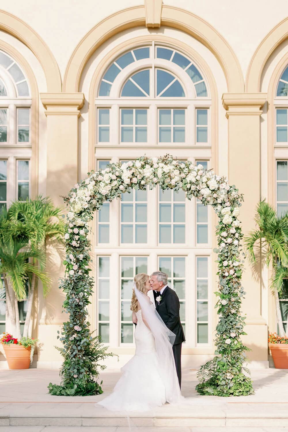 Beautiful wedding photo featuring a bride and groom under a floral arch in front of a grand building.