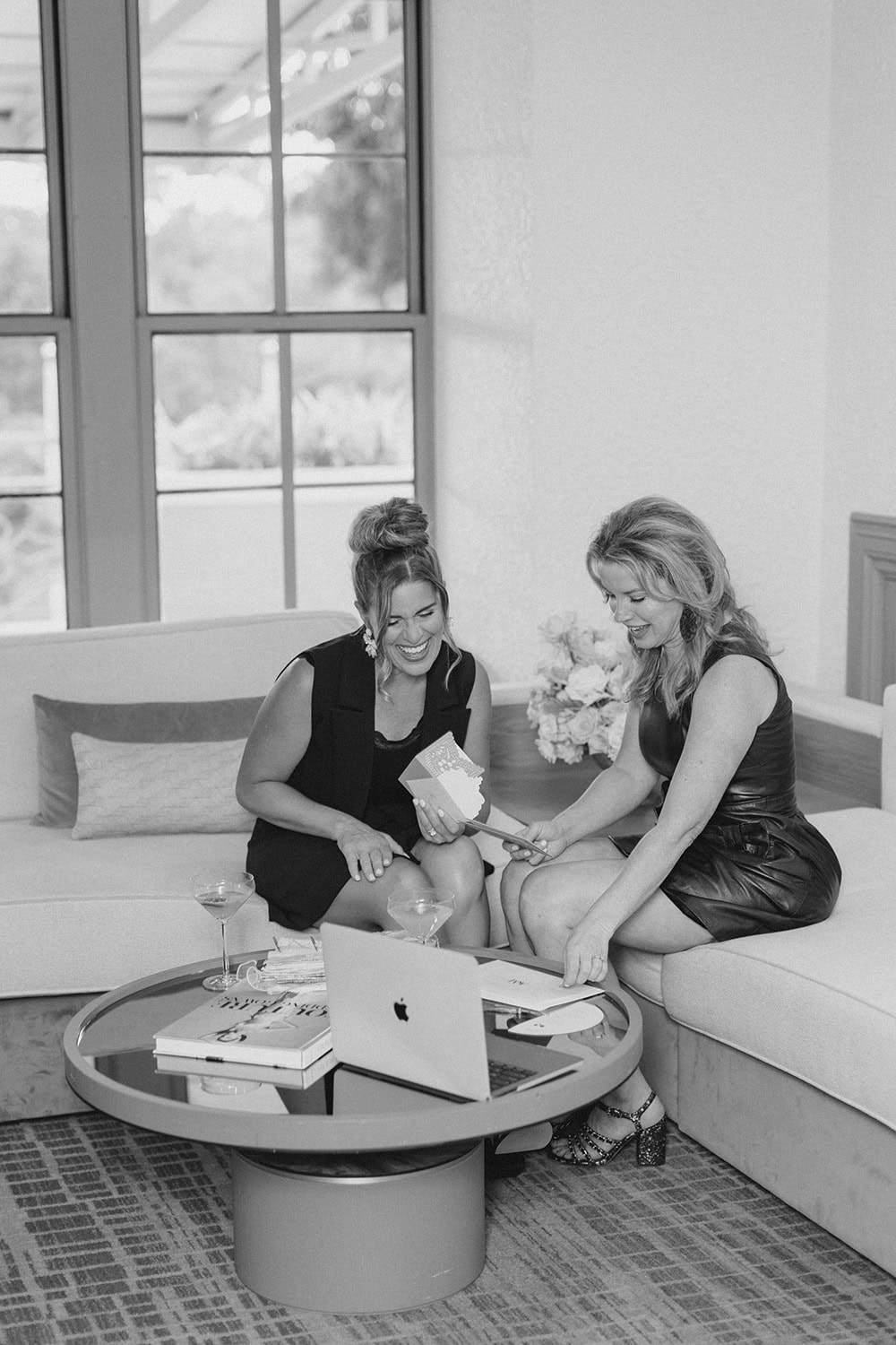Two women planning wedding details on sofa with laptop and flowers.