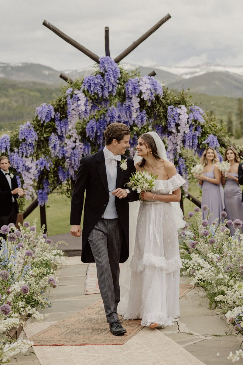 Bride and groom walking down aisle with floral arrangements and guests in background.