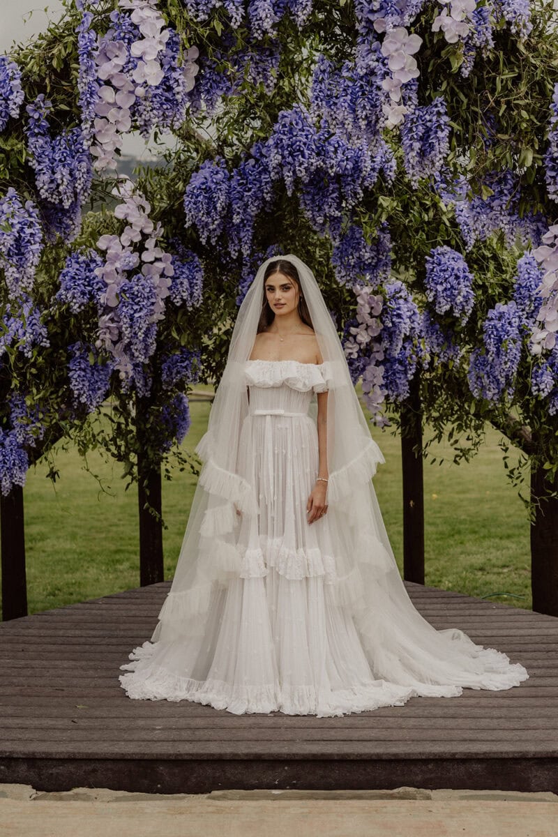 Bride in a white wedding dress standing on a wooden platform with purple and white flowers behind he.