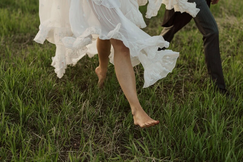 A woman in a flowing white dress and a man in dark striped pants dancing barefoot on grass.