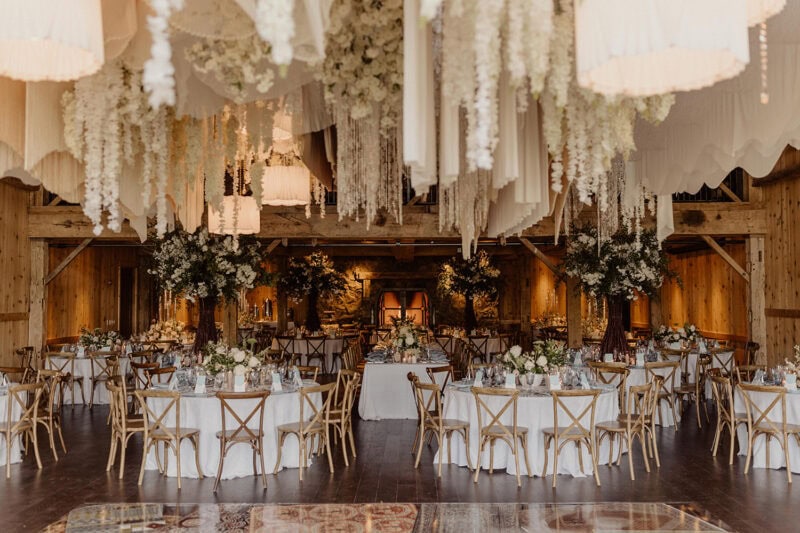 Beautiful wedding reception with floral centerpieces and hanging fabric decor in a rustic wooden ven.