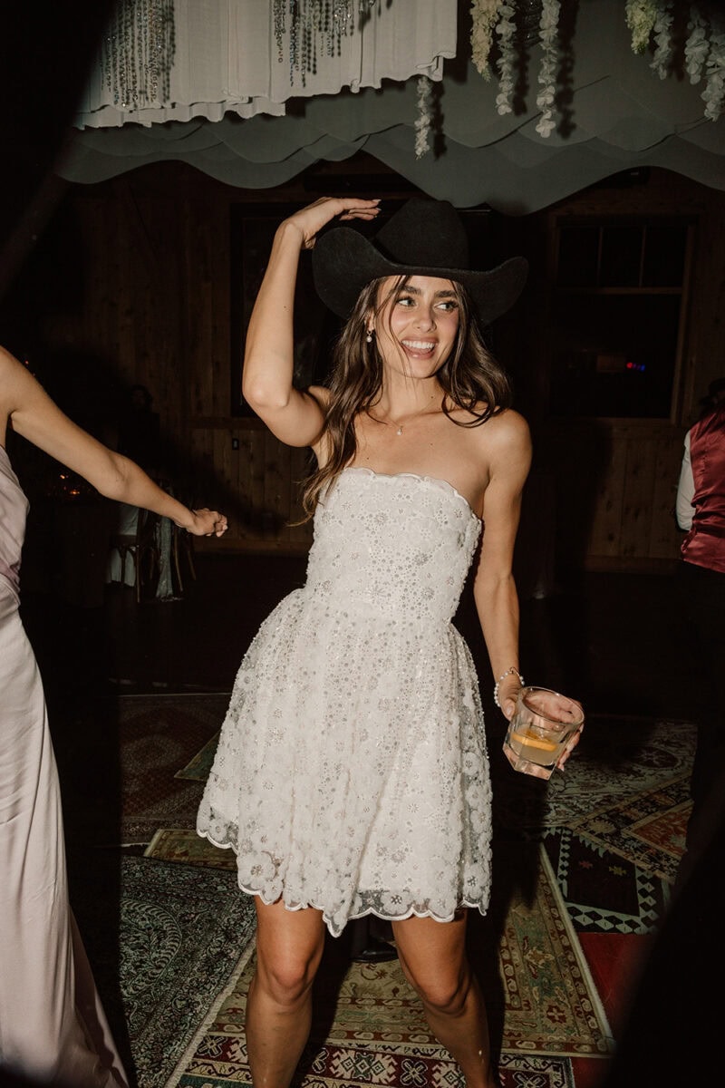 Happy bride wearing a white lace dress and a black cowboy hat at her wedding reception.