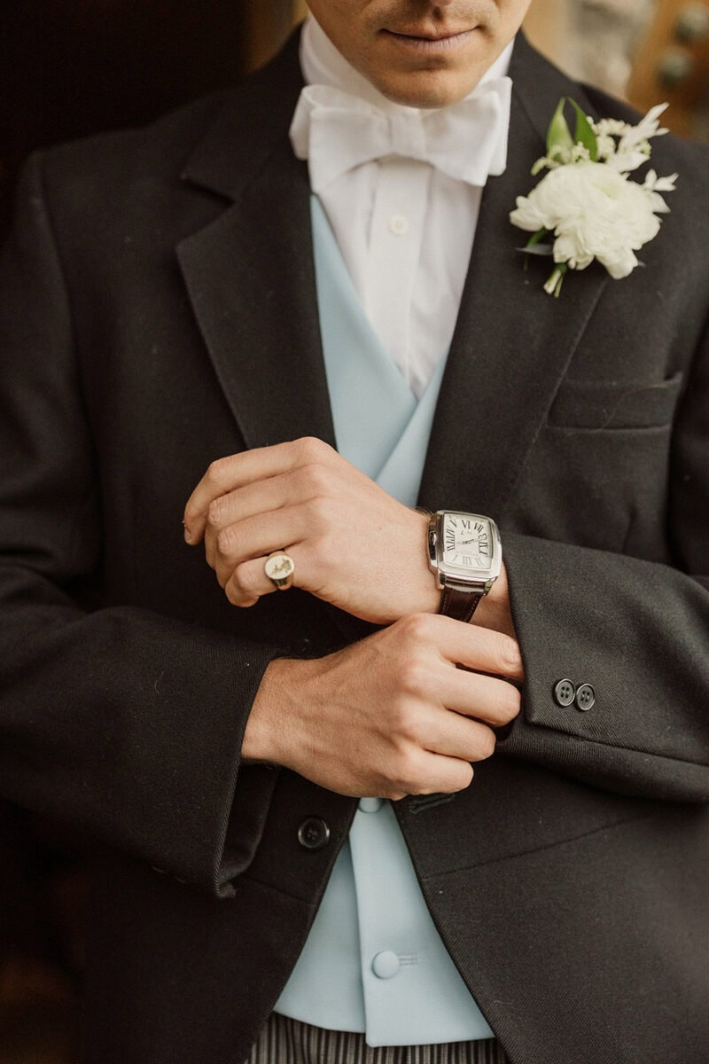 Groom in black tuxedo with white boutonniere, adjusting his watch before the wedding.