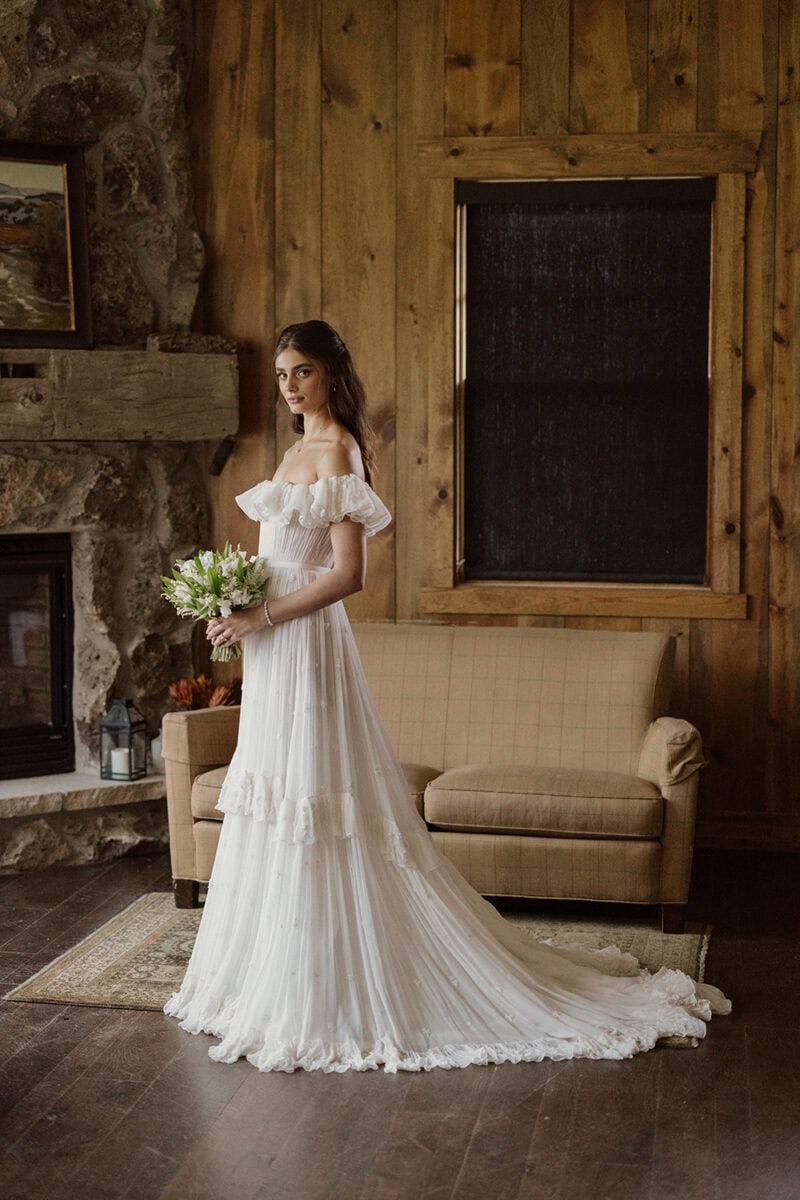 Bride in a white wedding gown holding a bouquet in a cozy rustic room setting.